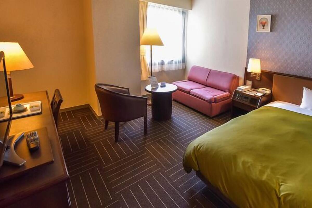 Quarto de hotel com uma cama, uma escrivaninha com luminária, uma cadeira, uma mesinha e um sofá rosa.