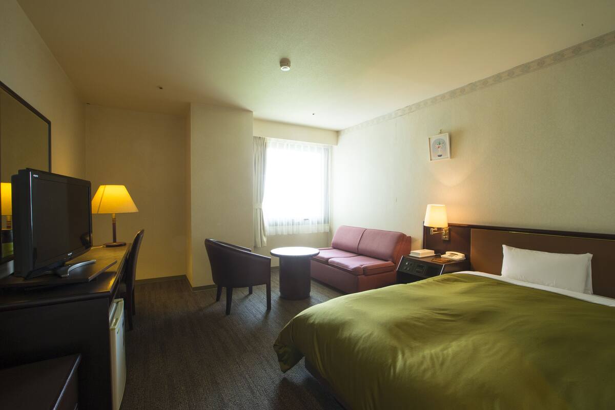 Quarto de hotel com cama, escrivaninha com computador, cadeira, sofá, mesinha e janela com cortinas.