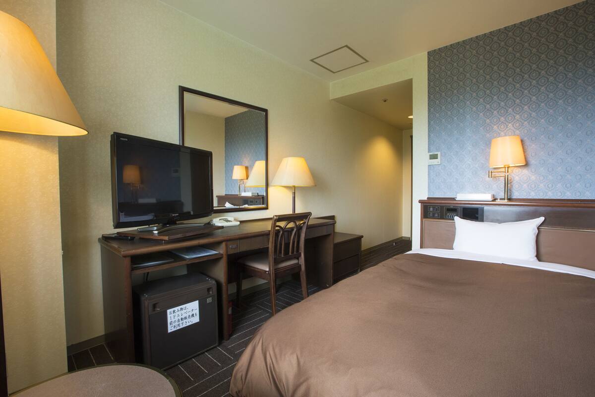Quarto de hotel com cama, escrivaninha, cadeira, televisão e luminárias.