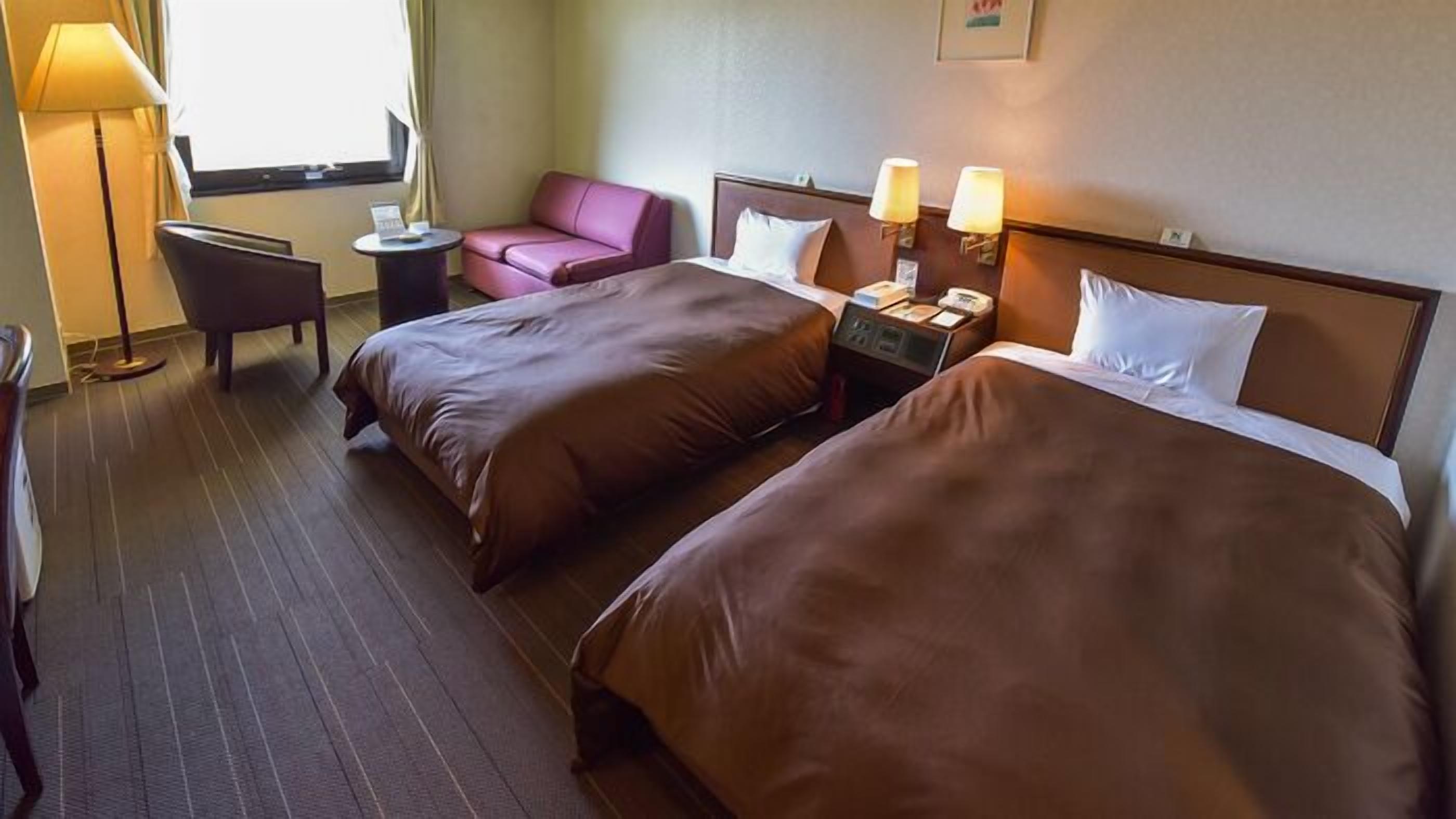 A hotel room with two beds, a desk, a chair, a lamp, and a window.