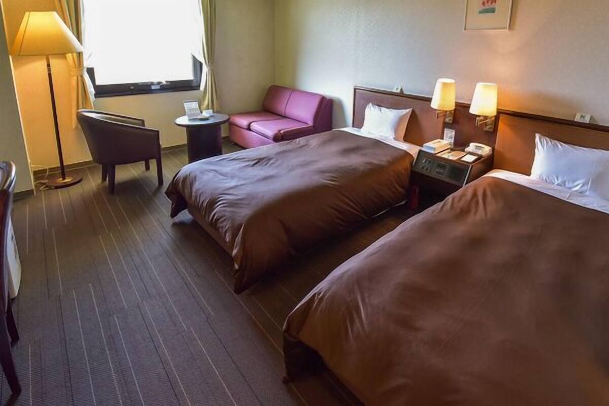 Quarto de hotel com duas camas, uma escrivaninha, uma cadeira, um abajur e uma janela.