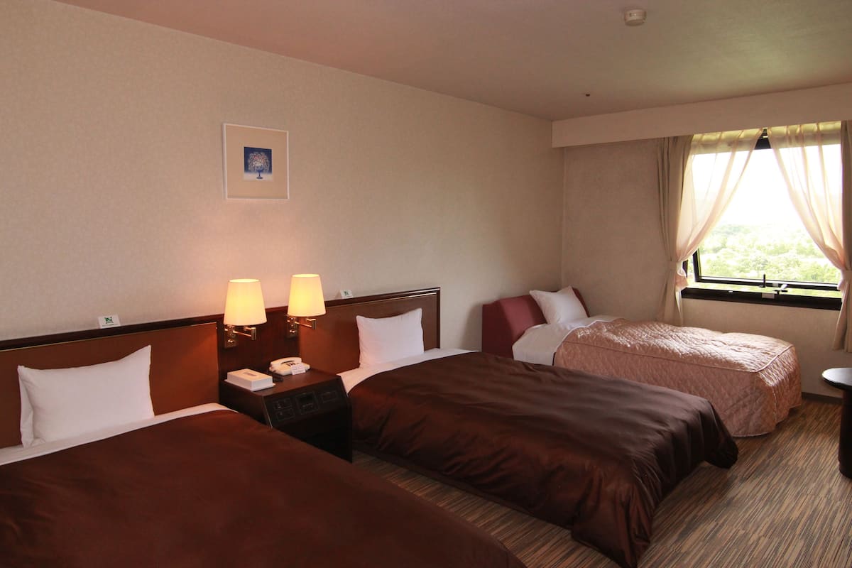 Quarto de hotel com duas camas, mesa de cabeceira com luminárias, janela com cortinas e uma pequena mesa.