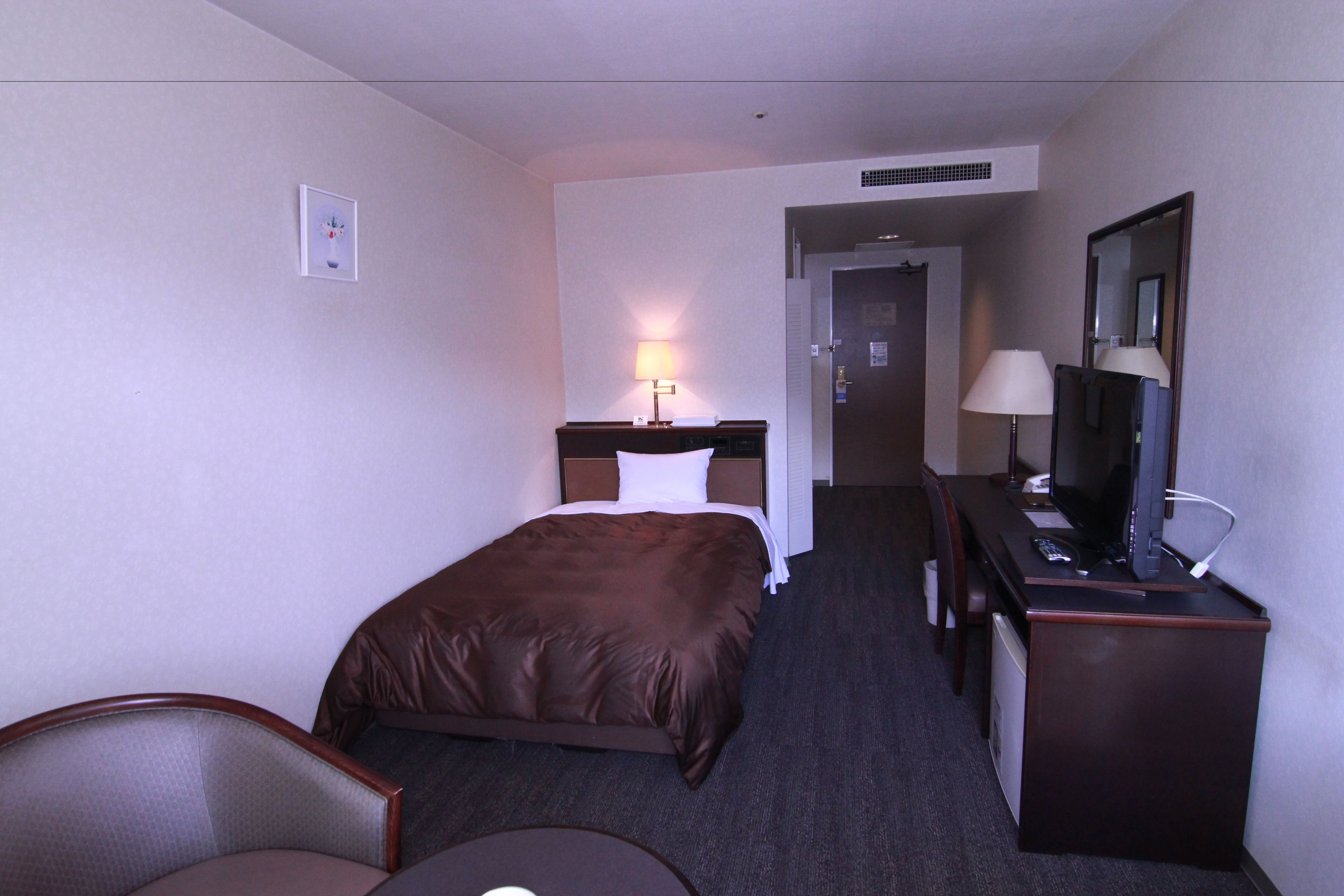 A hotel room with a bed, a desk with a computer, a chair, and a lamp.