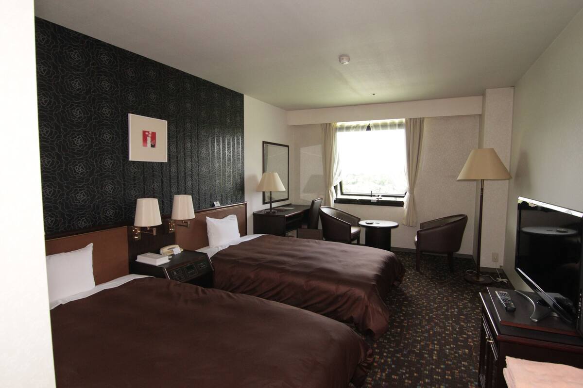 Quarto de hotel com duas camas, uma escrivaninha, uma cadeira, televisão e janela com cortinas.