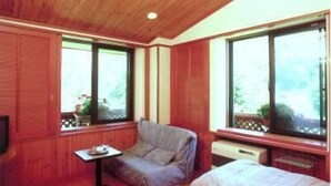 A cozy room with wooden paneling, a sofa, a small table, and large windows offering a view of greenery outside. - Hotel Gute Welle (Matsumoto)