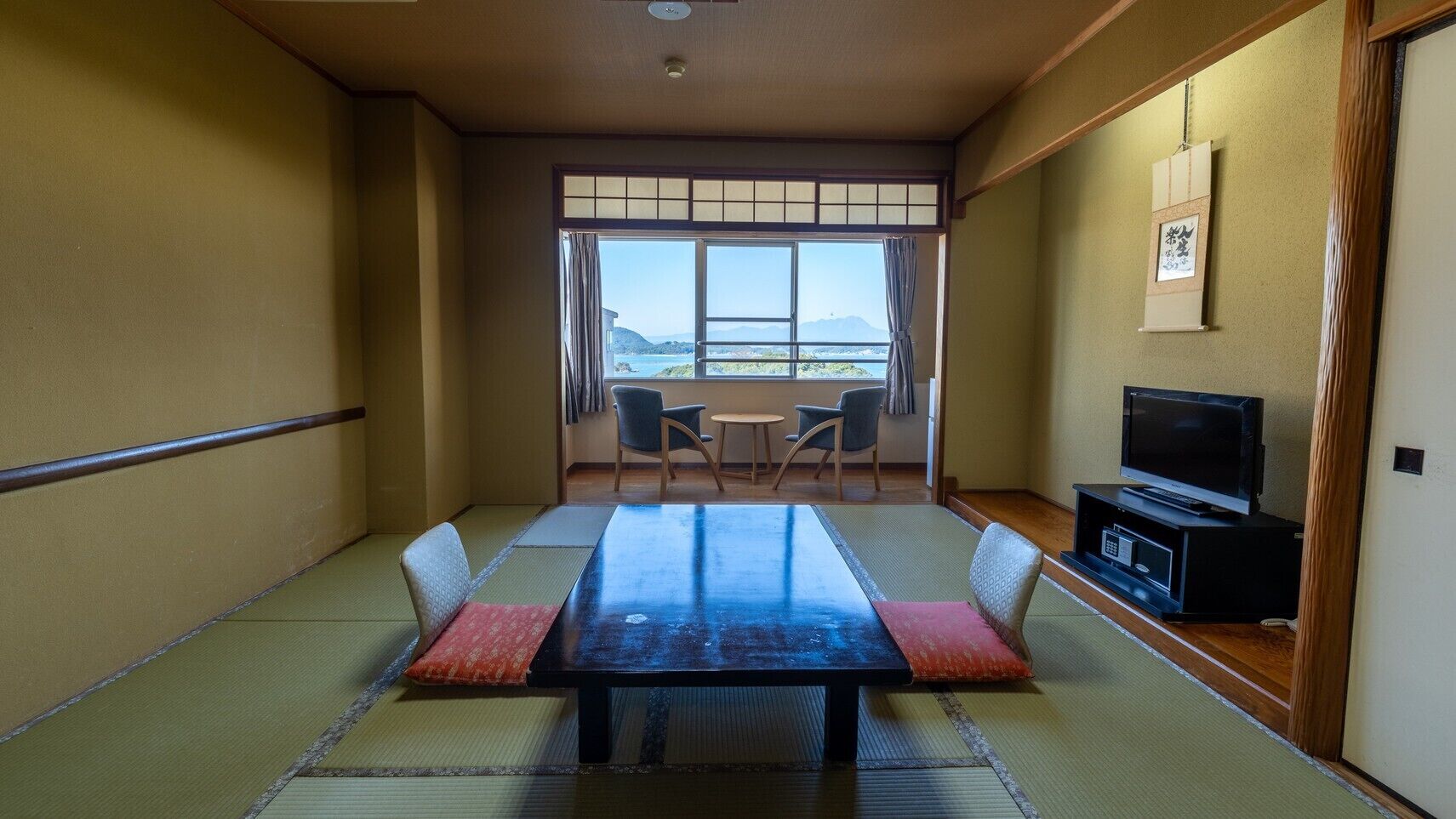 Ein traditioneller japanischer Raum mit Tatami-Böden, einem niedrigen Tisch, Stühlen, einem Fernseher und Blick ins Freie.