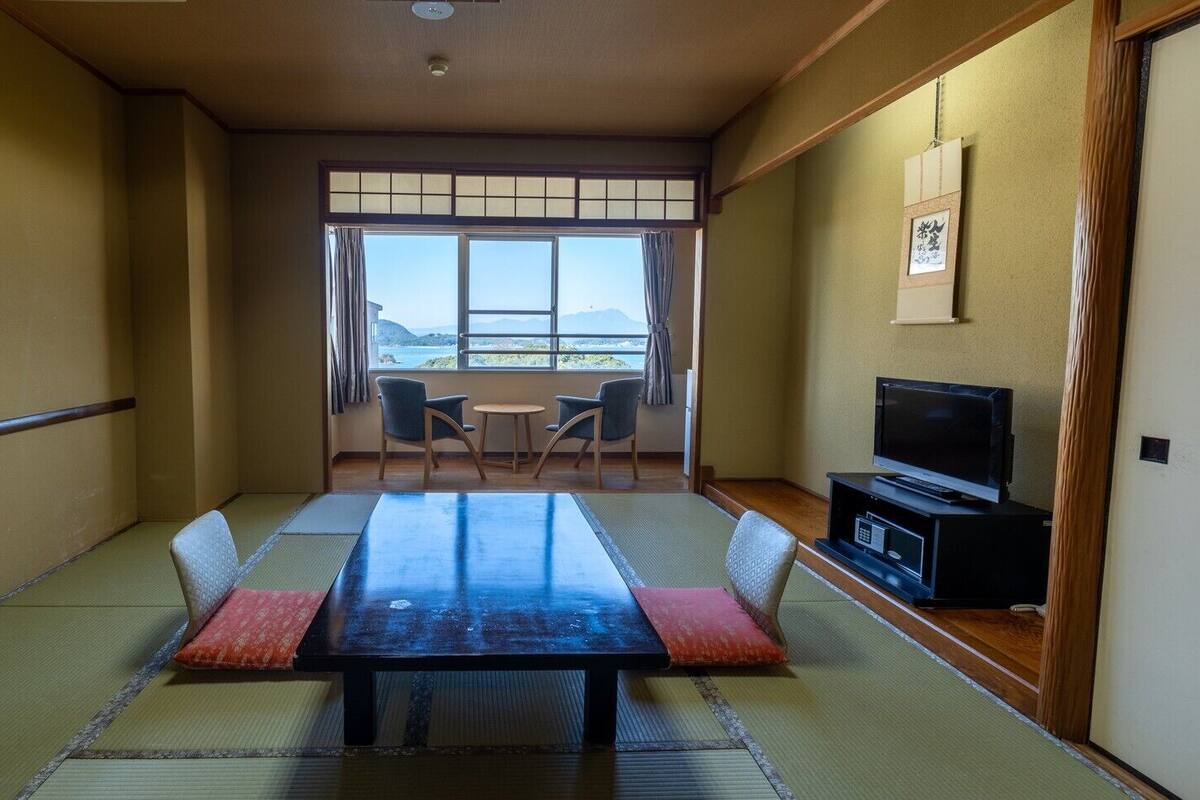A traditional Japanese room with tatami flooring, a low table, chairs, a TV, and a view of the outdoors.