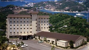 Un hotel con un edificio principal y una sección anexa más pequeña, rodeado de abundante vegetación y vistas al mar.