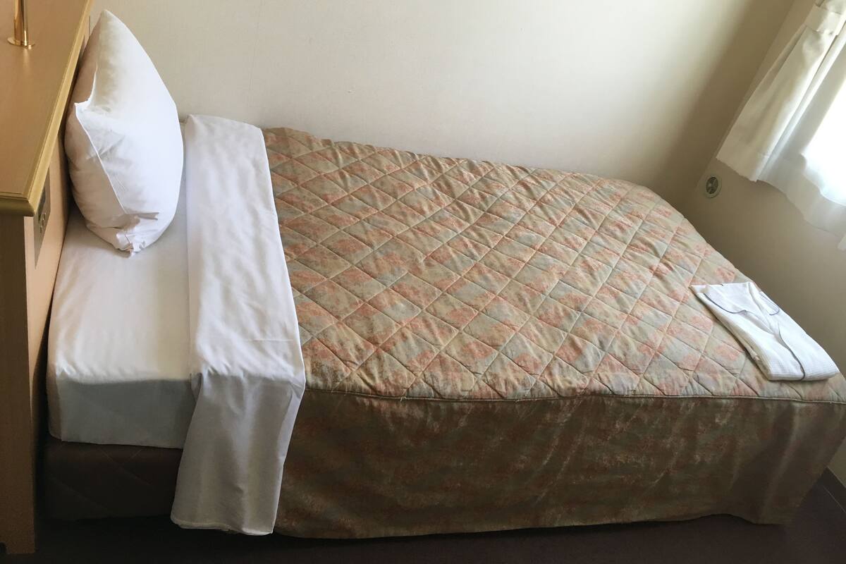 A single bed with a quilted cover, a white pillow, and a folded white garment on the bed.