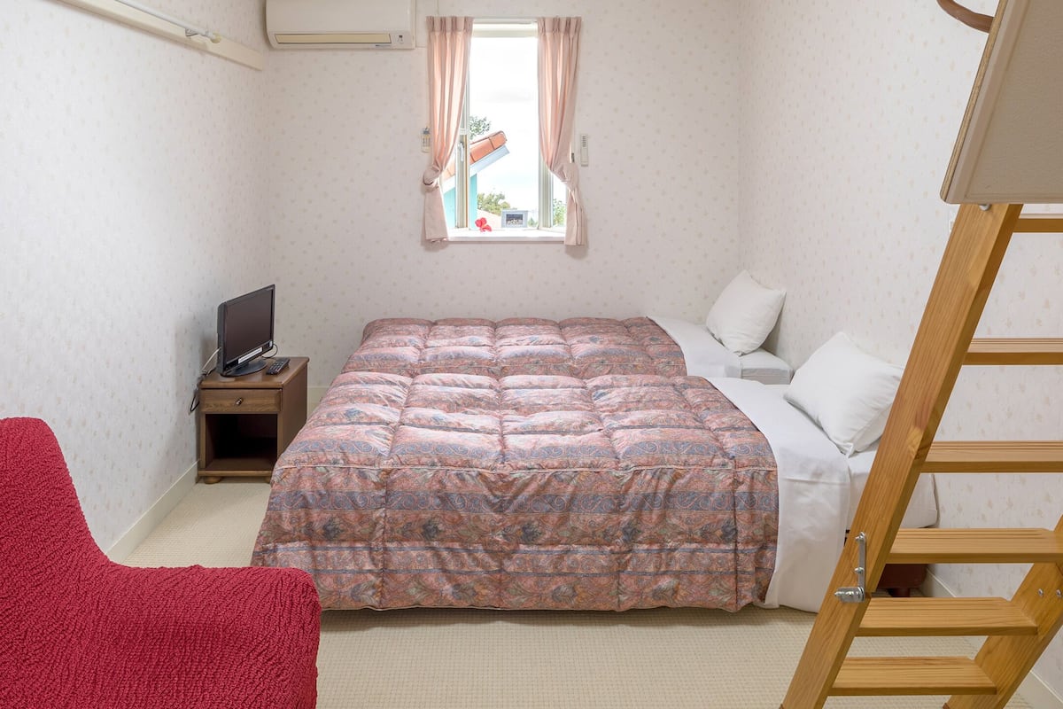 Ein Schlafzimmer mit einem Bett, einem Fernseher, einem roten Sessel, einem Fenster mit Vorhängen und einer Holzleiter.