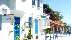 Un edificio blanco con contraventanas azules, un aro de baloncesto y un balcón con sillas azules y plantas en macetas.