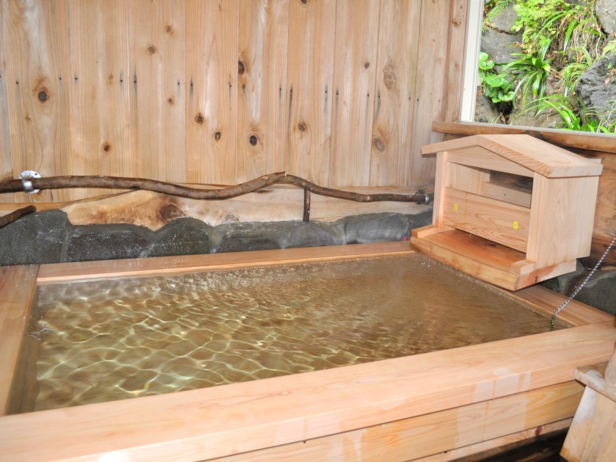 A wooden hot spring bath with a wooden bench and a small wooden structure, likely a changing room, against a backdrop of wooden panels and a stone wall.