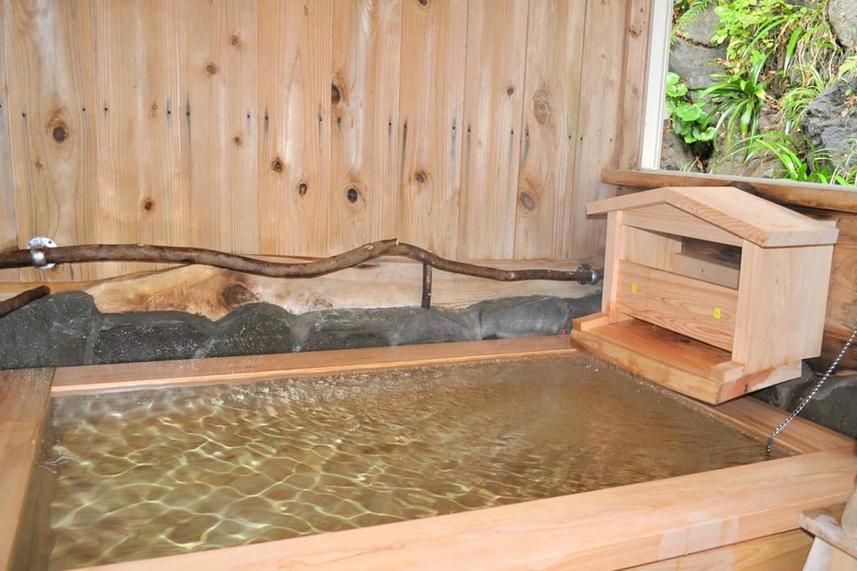 Ein hölzernes Quellbecken mit einer Holzbank und einer kleinen Holzstruktur, wahrscheinlich eine Umkleidekabine, vor einem Hintergrund aus Holzverkleidungen und einer Steinwand.