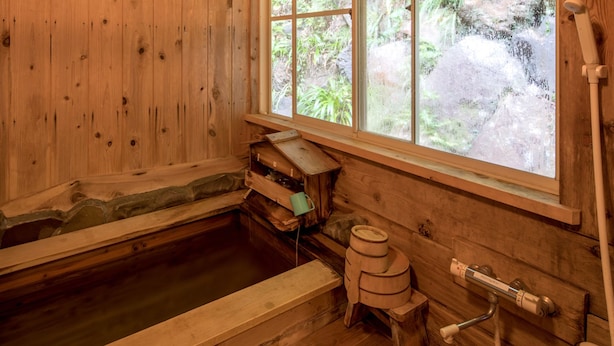 Habitación de madera con bañera de madera, una ventana que da a zonas verdes y un estante de madera con un objeto verde.
