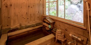A wooden room with a wooden bathtub, a window with a view of greenery, and a wooden shelf with a green object. - Mediterranean Sea Pension Knossos (Higashiizu)