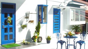 Un edificio blanco con puertas y ventanas azules, un pequeño espacio al aire libre con sillas metálicas azules y plantas en macetas.