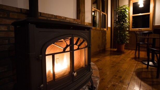 A wood-burning stove with a glass door, a brick wall, wooden flooring, a potted plant, and a window with a hanging light fixture. - BLISS Villa TSUBAKI (Kutchan)