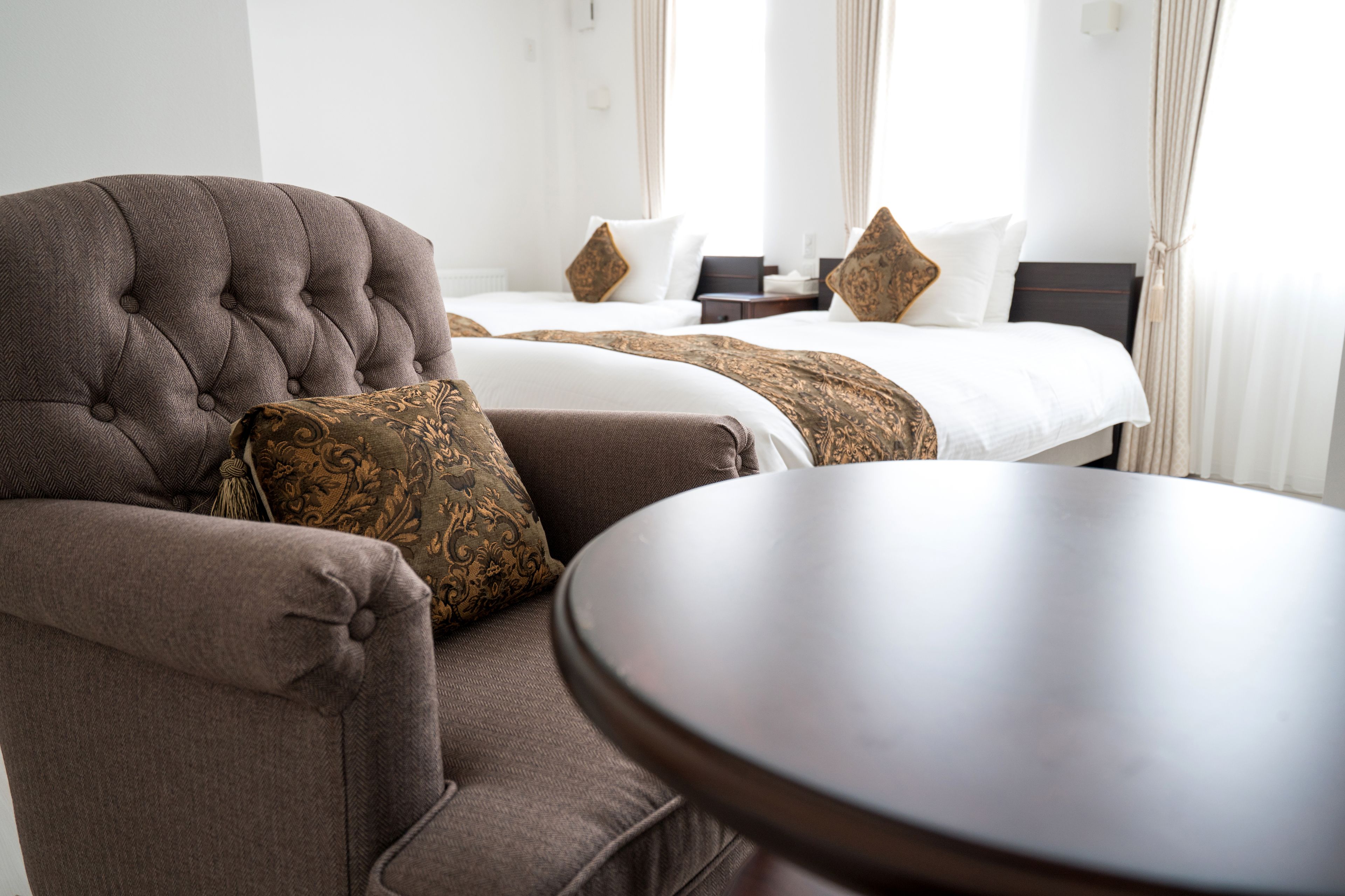 A hotel room with a tufted armchair, a round table, and two beds with white bedding and patterned throw pillows.