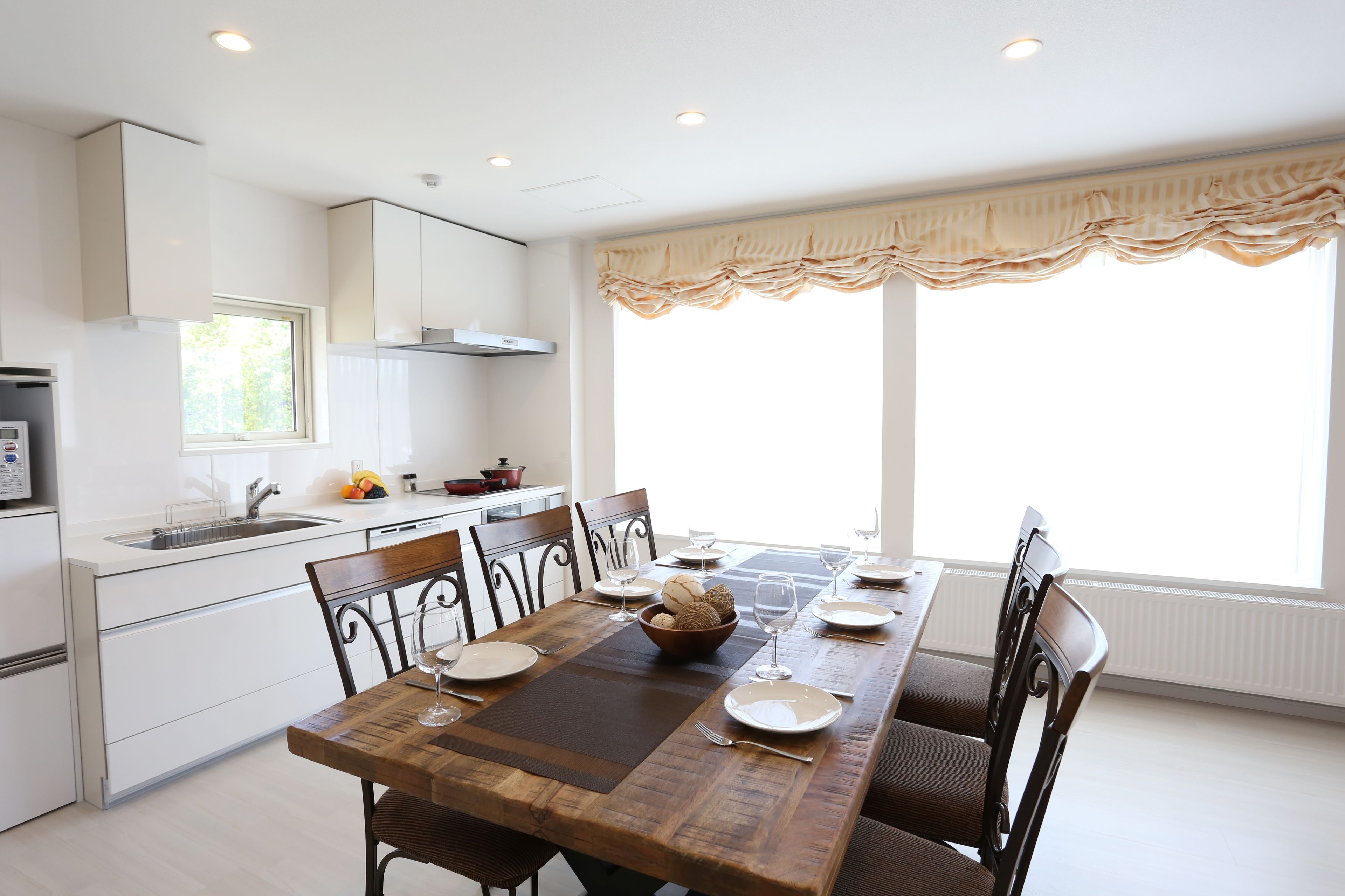 A modern kitchen with white cabinets, a wooden dining table set for four, and a large window with a valance.