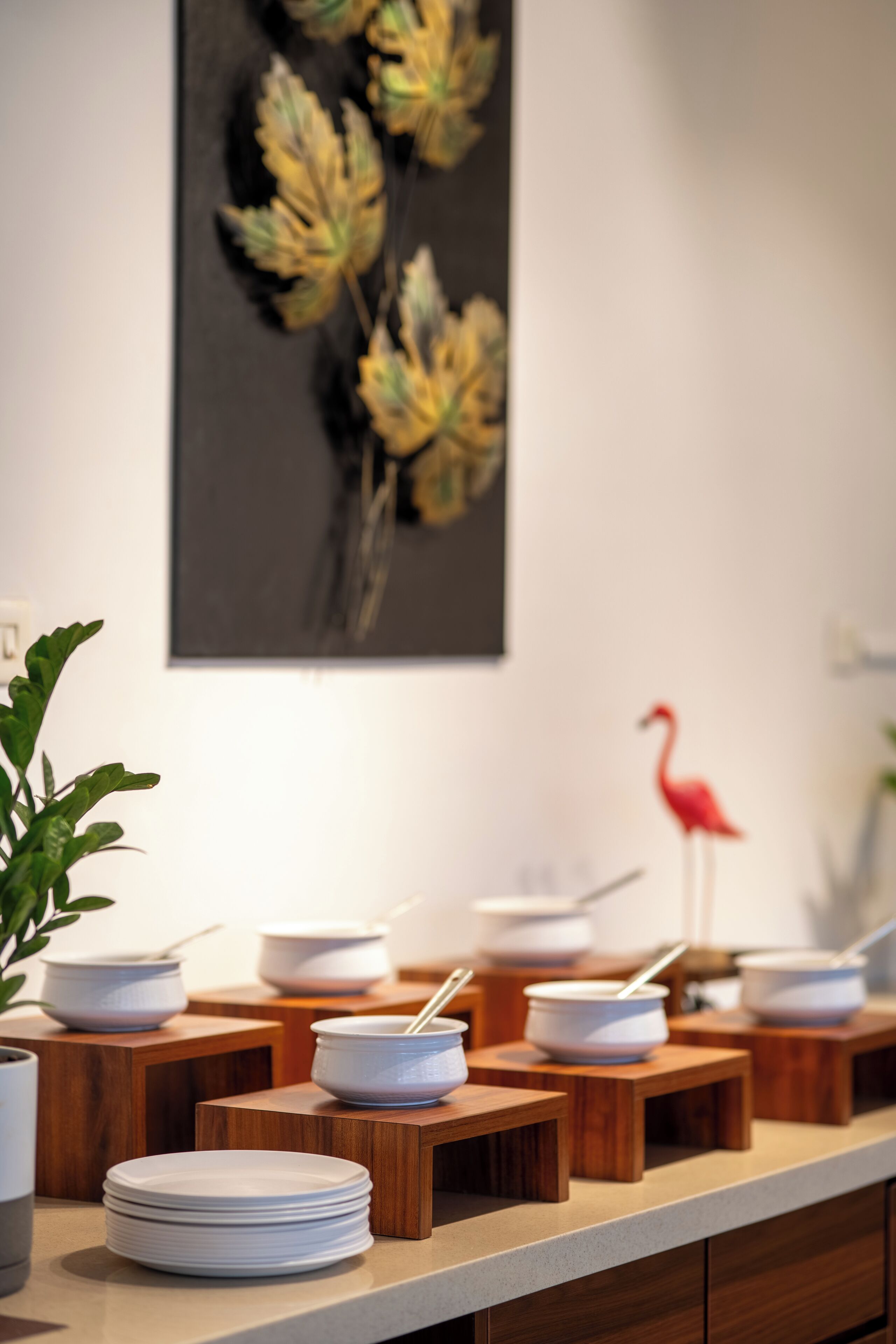 A dining area with multiple white bowls on wooden stands, a red flamingo decoration, and a painting of leaves on the wall.