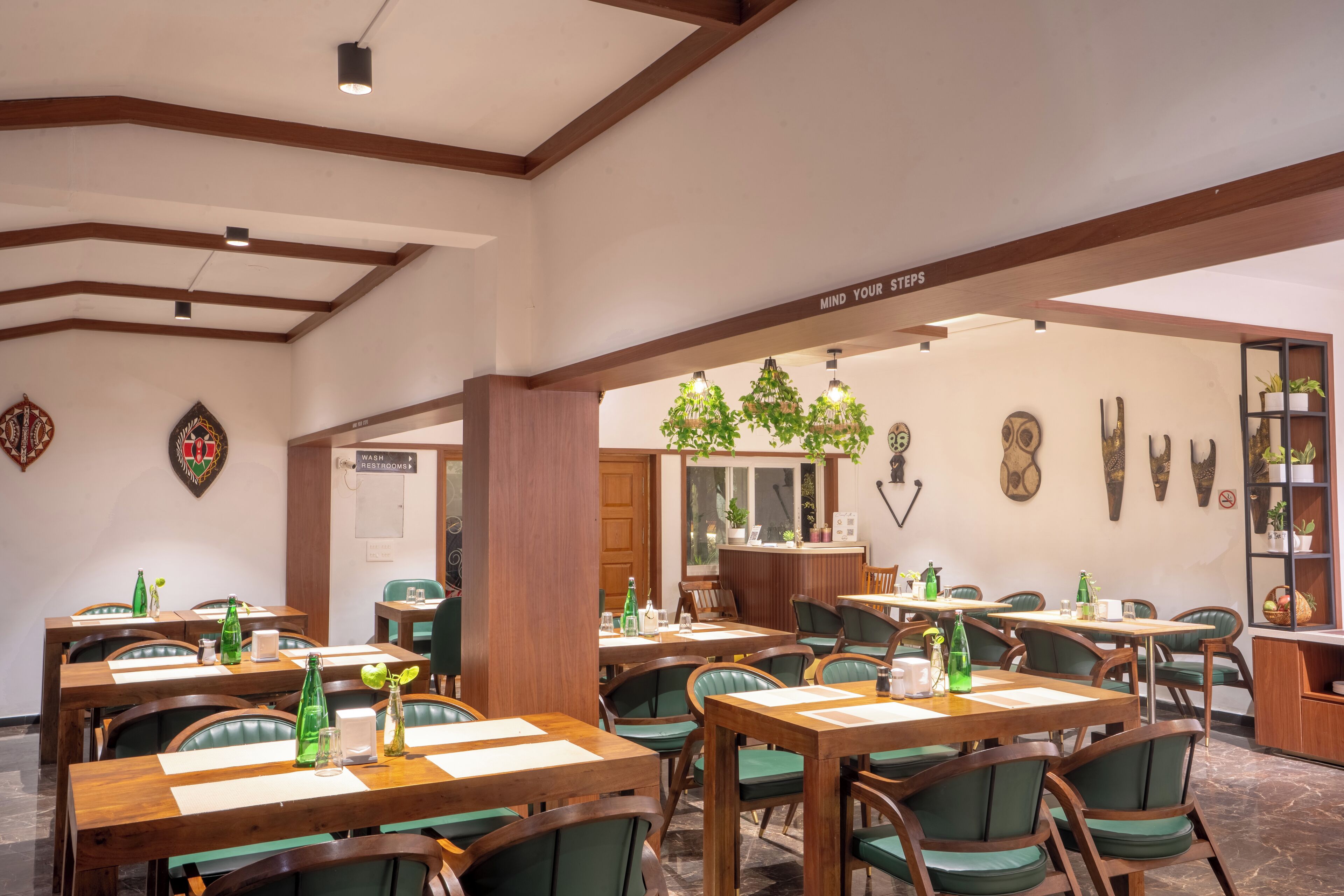 A restaurant with wooden tables and green chairs, a sign reading "MIND YOUR STEPS", and various wall decorations including a clock and framed items.