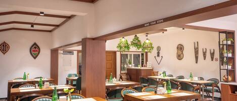 A restaurant with wooden tables and green chairs, a sign reading "MIND YOUR STEPS", and various wall decorations including a clock and framed items.