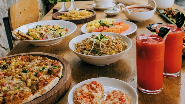 A table set with various dishes, including pizza, salads, and drinks, in an outdoor setting with a stone wall and greenery in the background.