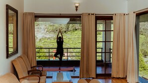 A room with a large sliding glass door leading to a balcony with a view of a green landscape. There is a yoga mat on the balcony, and a person is doing yoga.