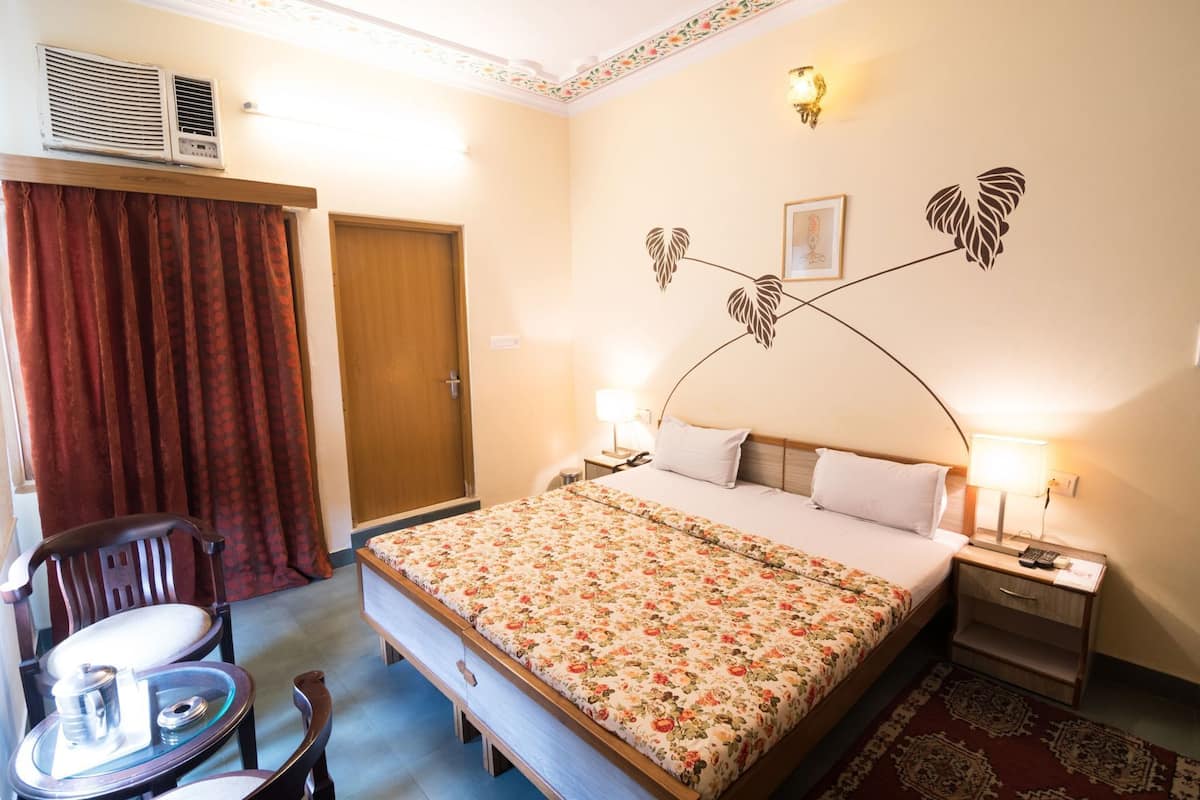 Habitación de hotel con cama, mesitas de noche, una silla, una mesita pequeña, ventana con cortinas y aire acondicionado de pared.