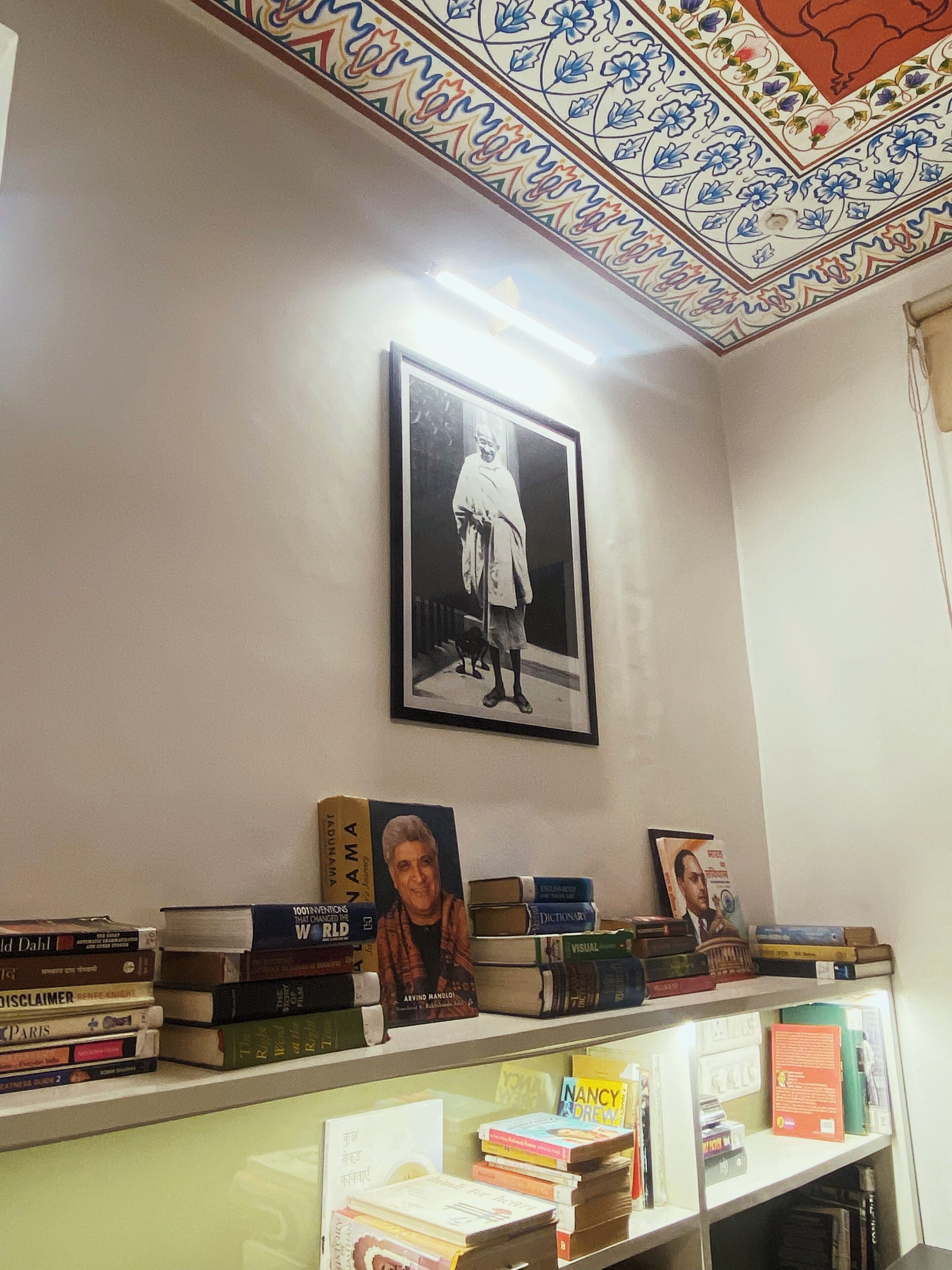 A room with a bookshelf filled with books, a framed photograph on the wall, and a decorative ceiling.
