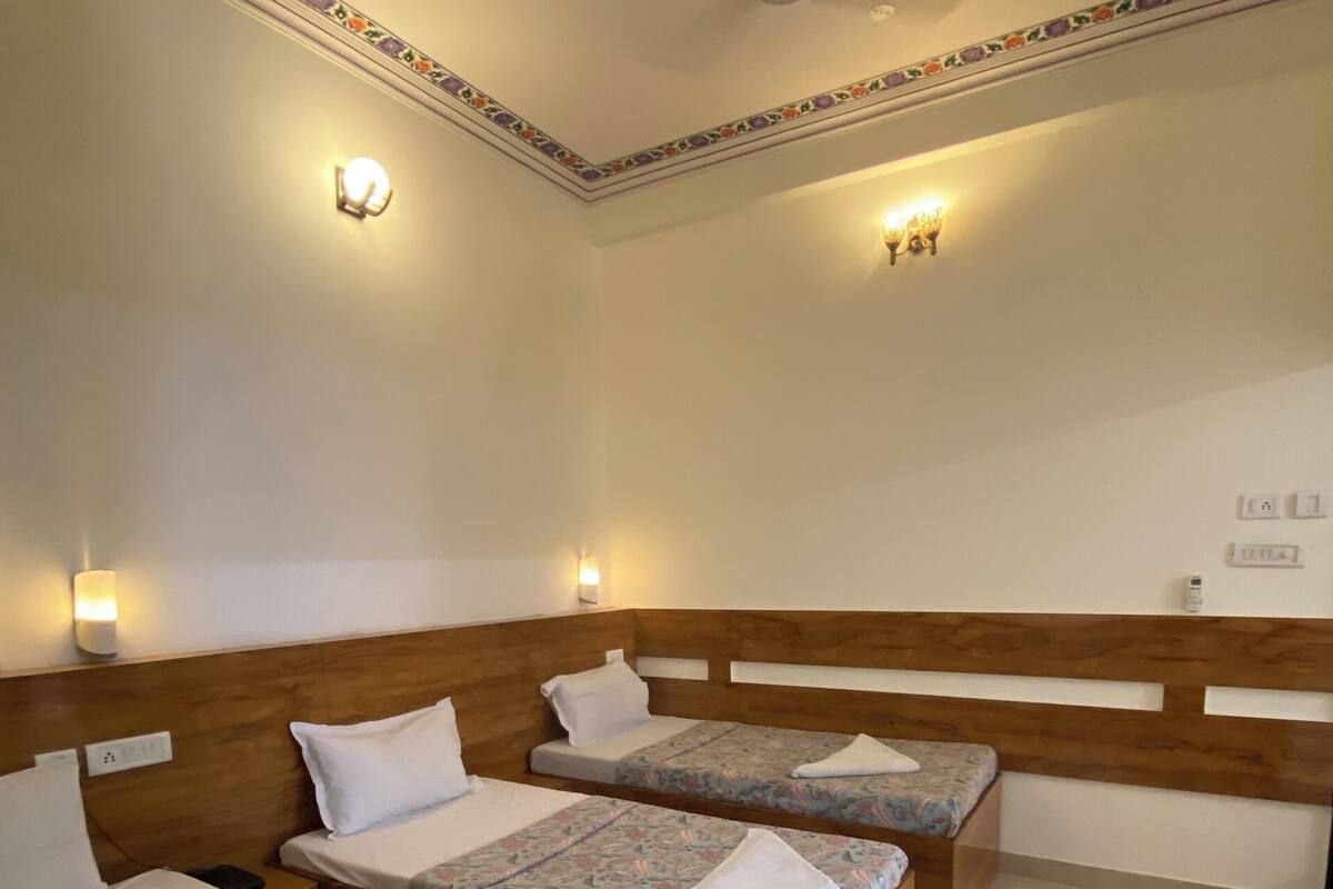 Habitación de hotel con tres camas individuales, cada una con cabecero de madera y ropa de cama estampada. Hay luces fijadas a la pared y un ventilador de techo.