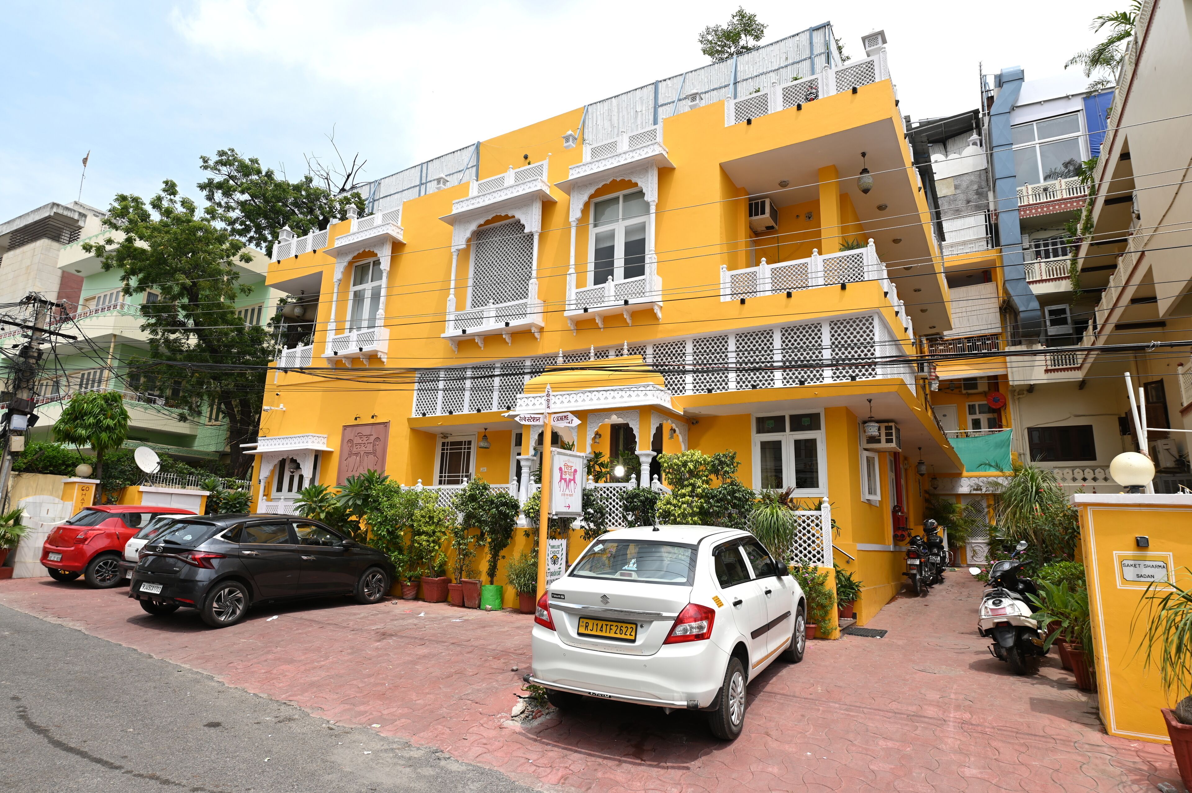 Un edificio amarillo de varios pisos con balcones, coches aparcados y plantas en macetas.
