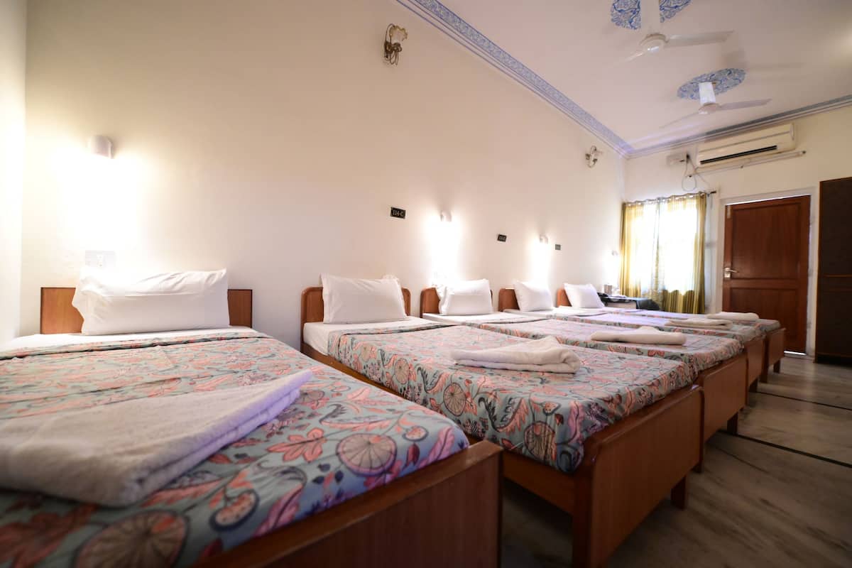 Habitación de hotel con cuatro camas individuales, cada una con una colcha de estampado floral y almohadas blancas. Hay luces fijadas a la pared sobre cada cama y un equipo de aire acondicionado en la pared.