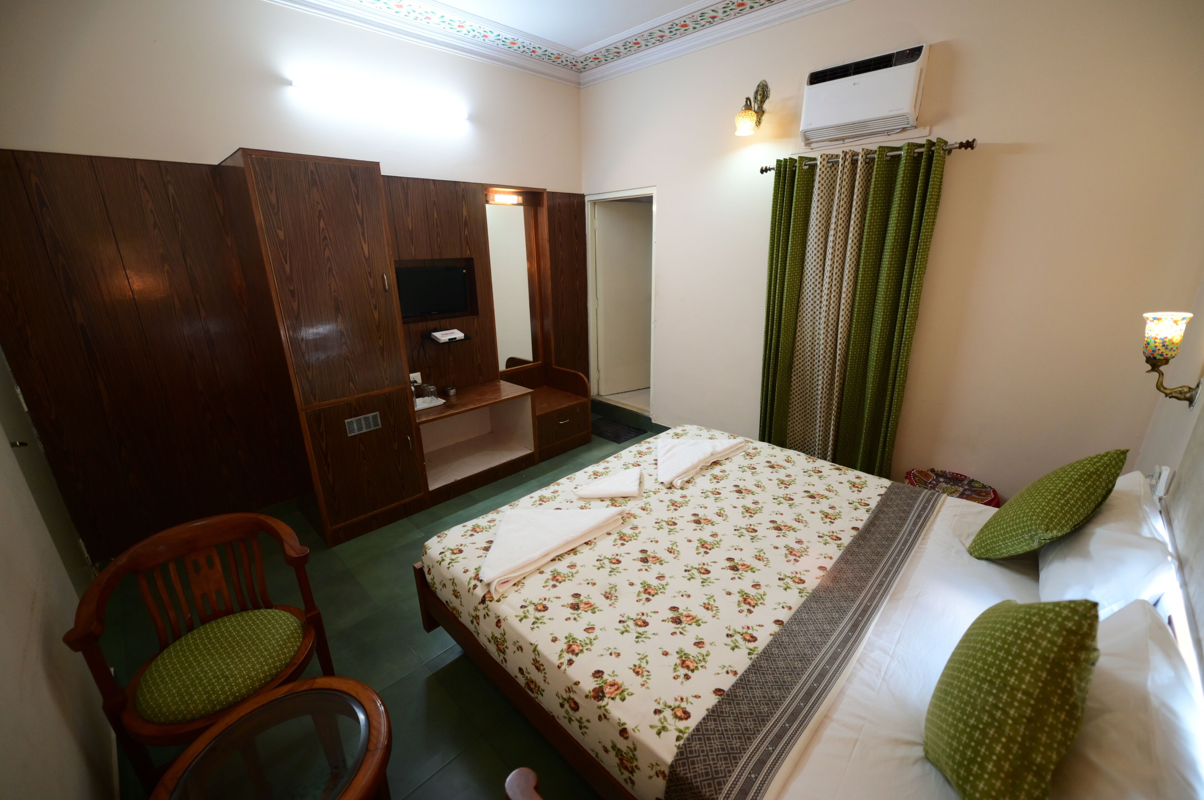 A hotel room with a bed, a wooden wardrobe, a desk with a chair, a television, and an air conditioning unit.