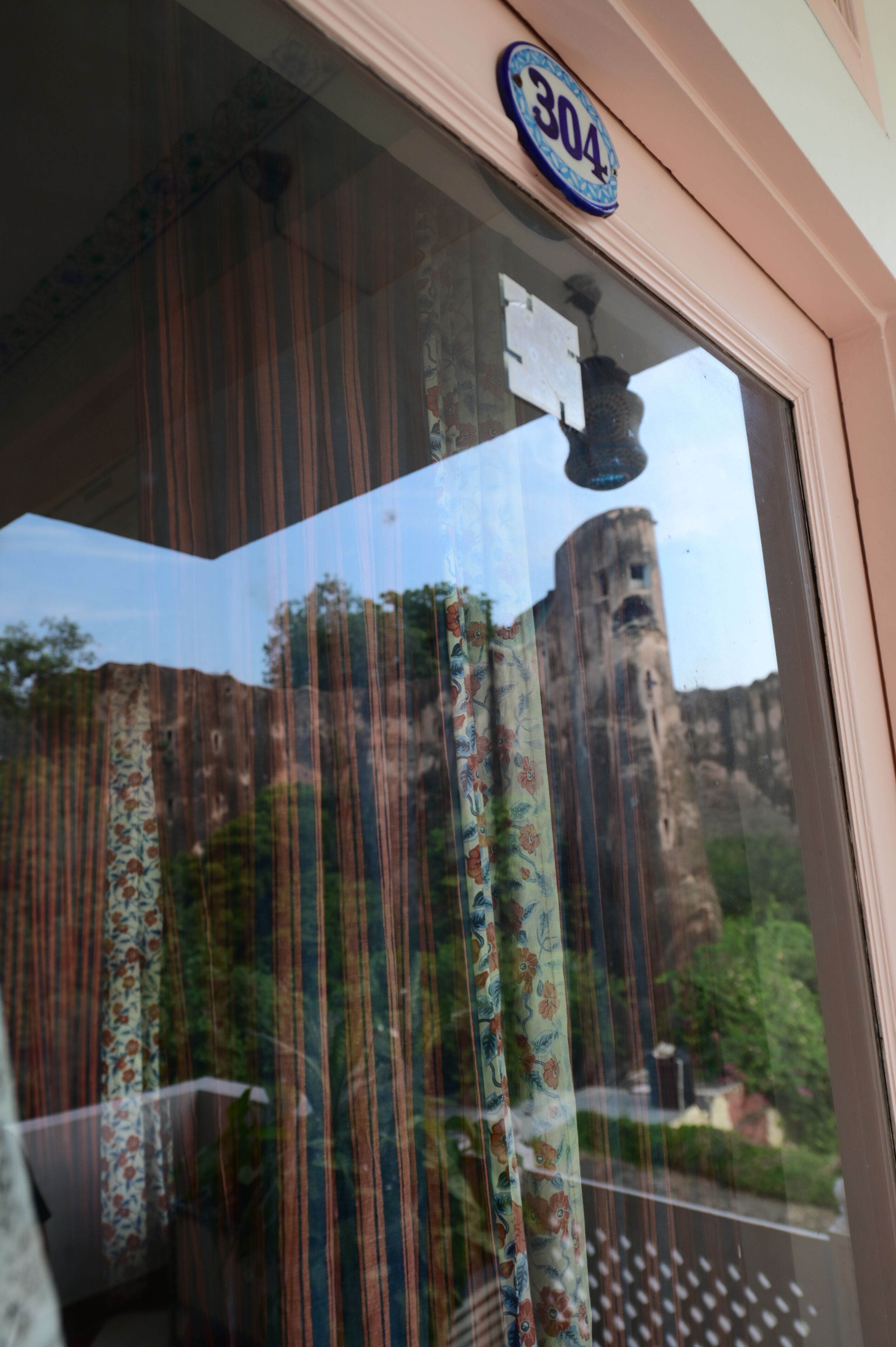 A window with a floral curtain, a number sign '304', and a view of a building and greenery outside.