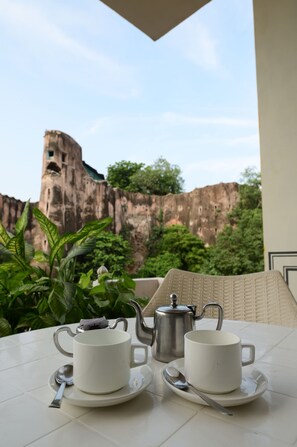 Une table avec deux tasses et sous-tasses blanches, une théière et une cuillère, le tout placé devant un décor de verdure et une falaise rocheuse.