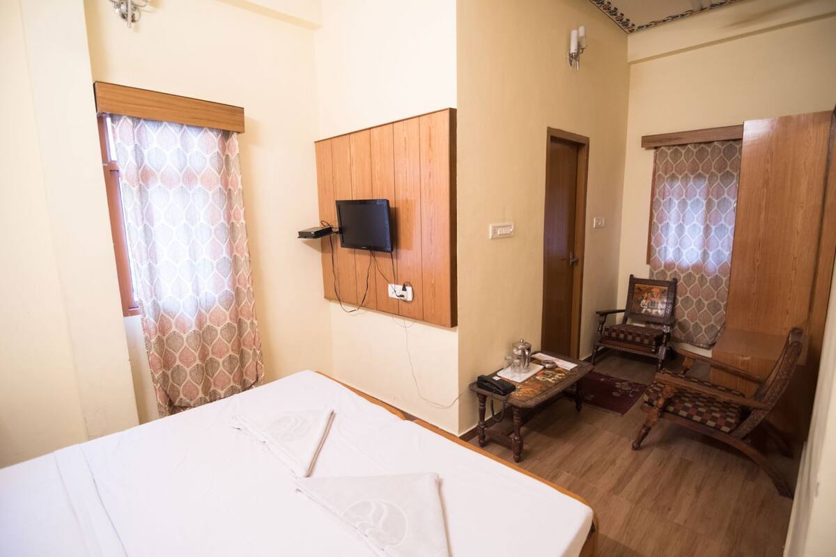 Una habitación con una cama, un televisor en la pared, un armario de madera, una mesita de noche con un marco de fotos y una cortina estampada.
