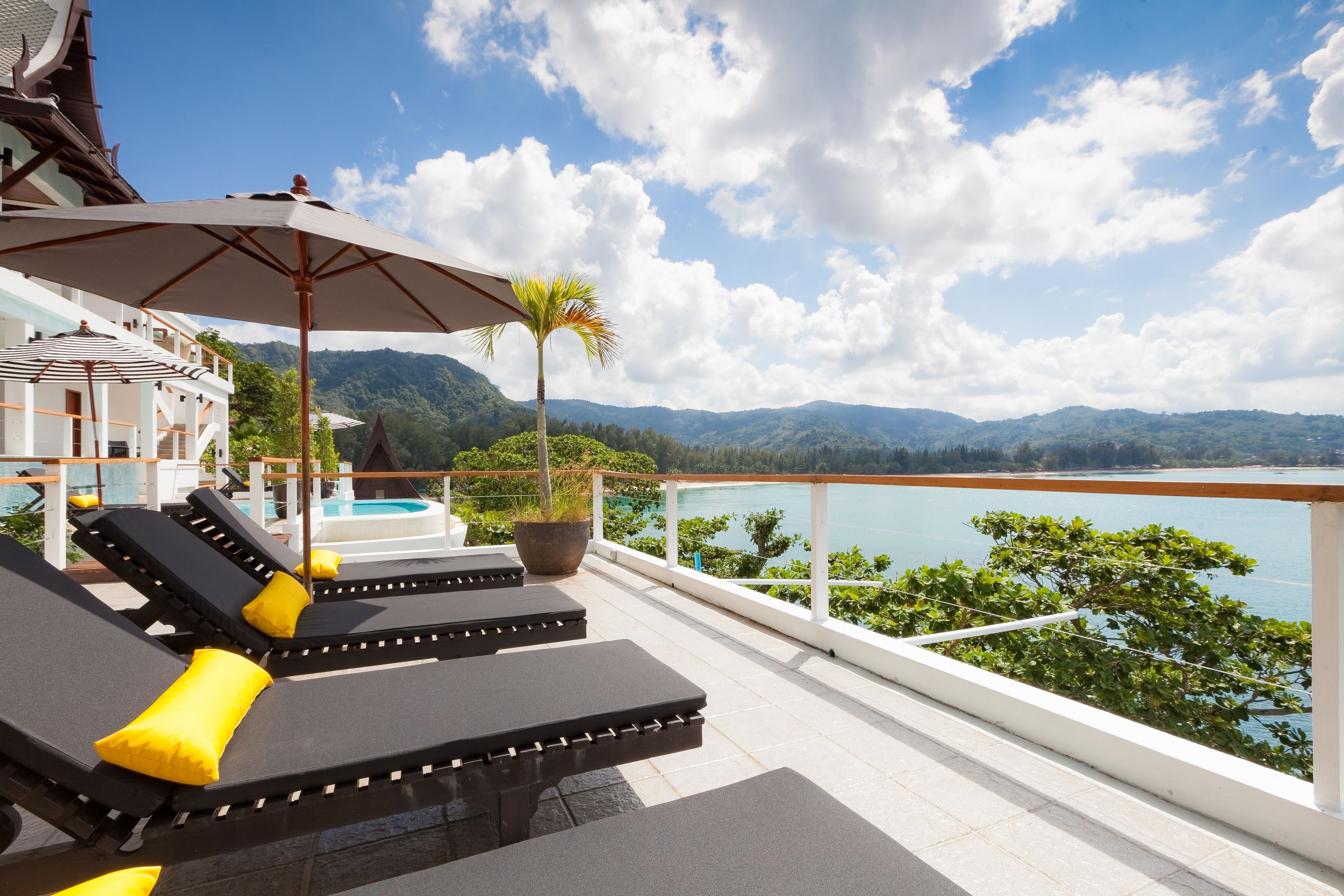 A balcony with sun loungers and a large umbrella, overlooking a body of water and mountains.
