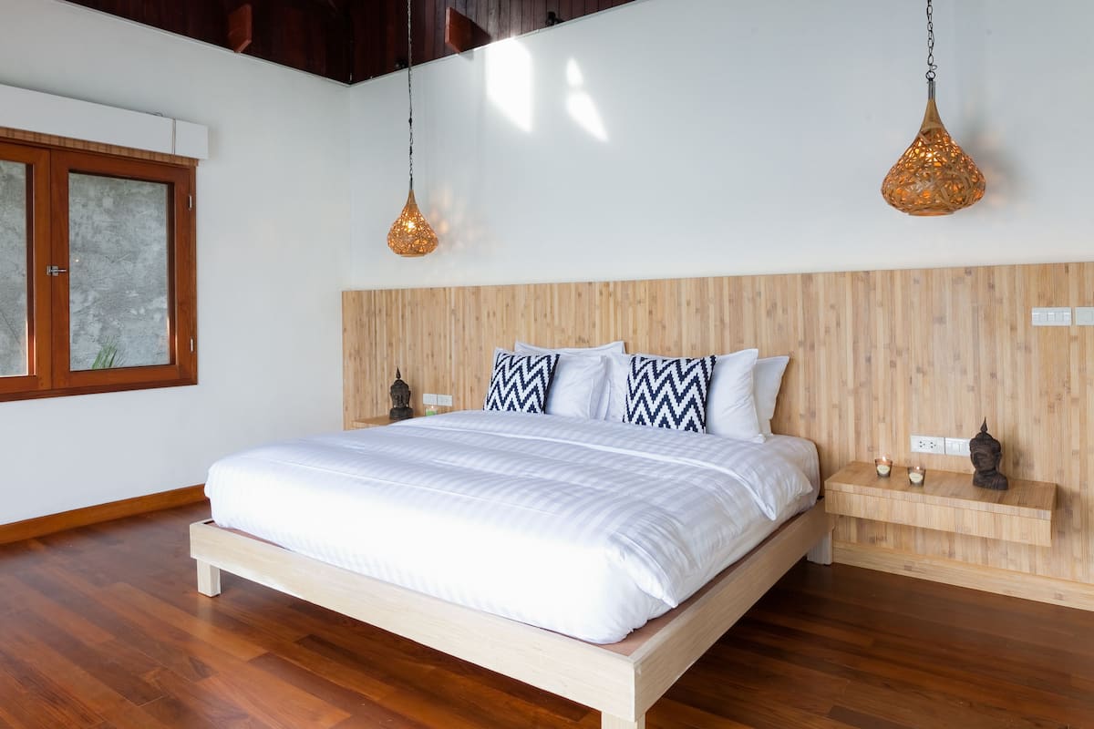 Un dormitorio con cama de madera, dos lámparas colgantes, una ventana con vidrio esmerilado y una mesita de noche de madera con una estatua de Buda.