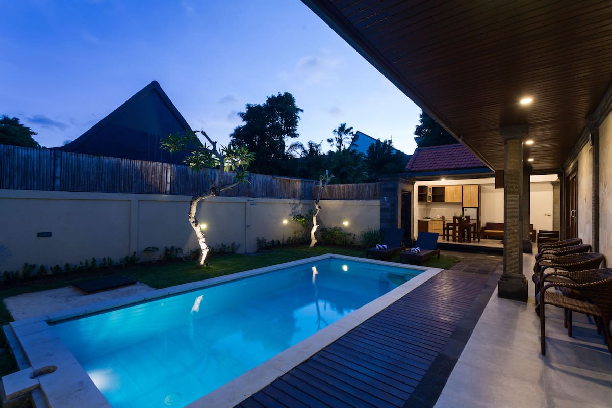 A swimming pool with a wooden deck, a covered patio area with chairs, and a view of the backyard.