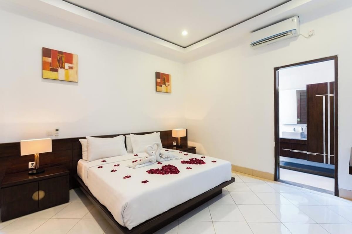 A hotel room with a large bed, two bedside tables, a nightstand, a wall-mounted air conditioner, and two paintings on the wall.