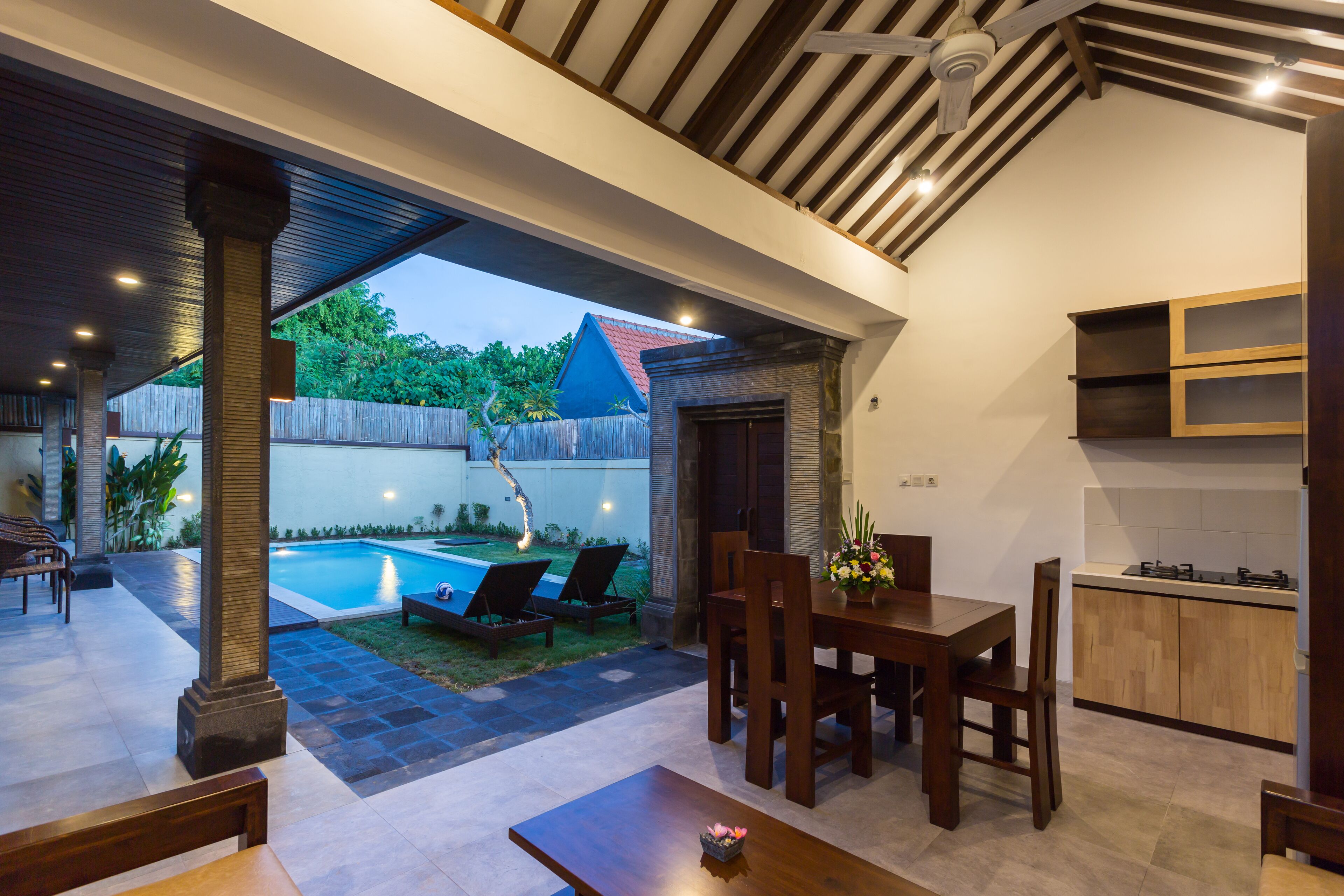 A covered outdoor dining area with a pool, wooden furniture, and a kitchen area.