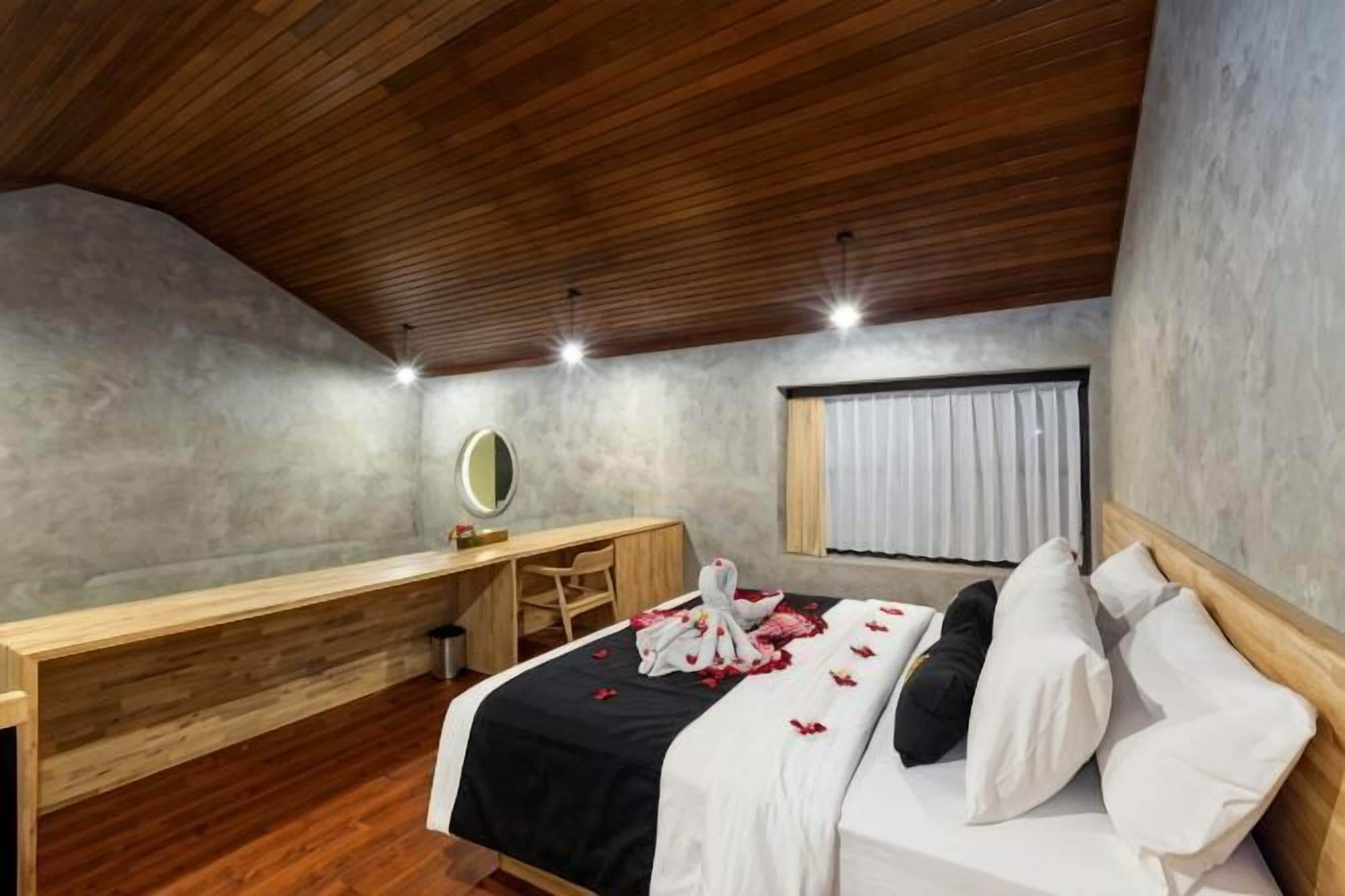 A bedroom with a wooden ceiling, a desk, a bed with white bedding, and a window with curtains.