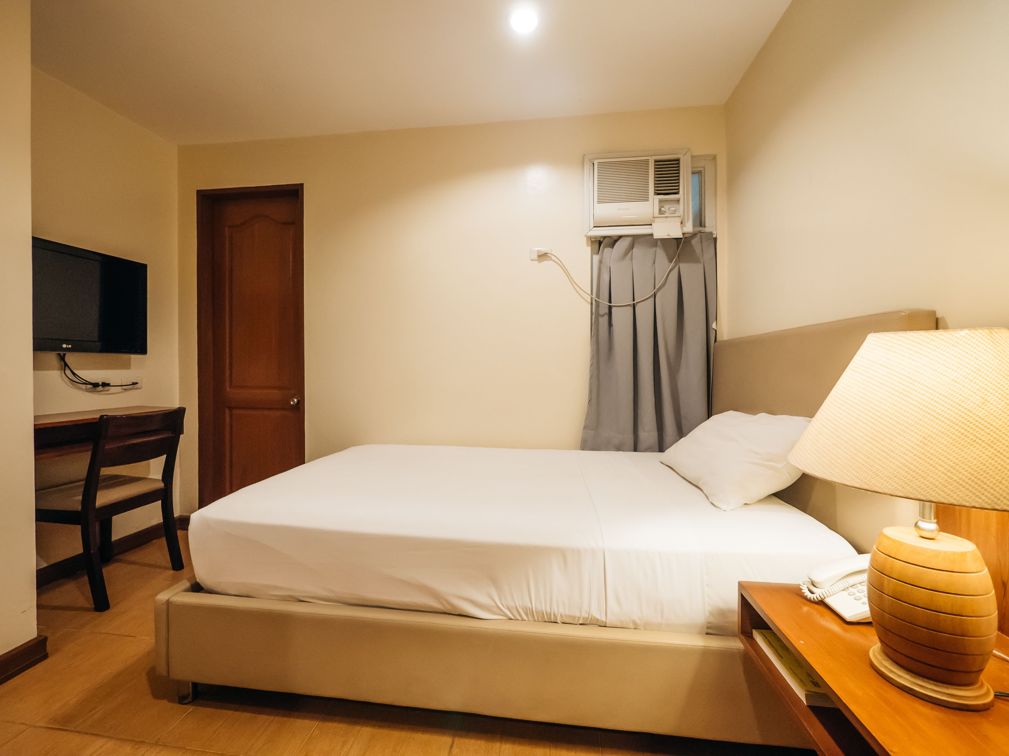 Habitación de hotel con una cama, un escritorio con televisor, un equipo de aire acondicionado, una lámpara y un teléfono.