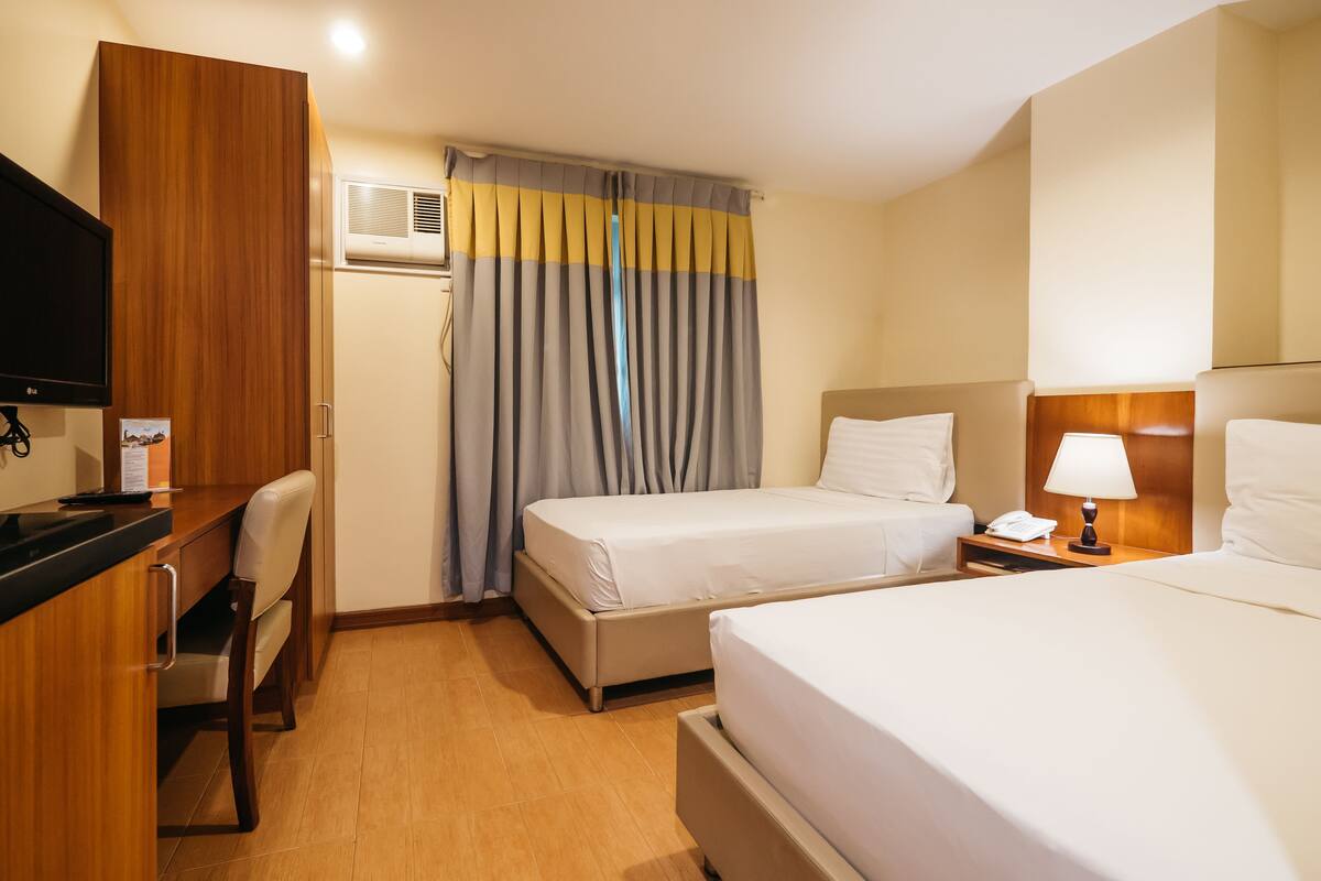 A hotel room with two beds, a desk with a chair, a TV, and a bedside table with a lamp.