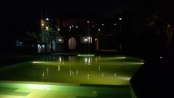A nighttime view of a swimming pool area with a fountain, surrounded by a low wall and illuminated by ambient lighting.