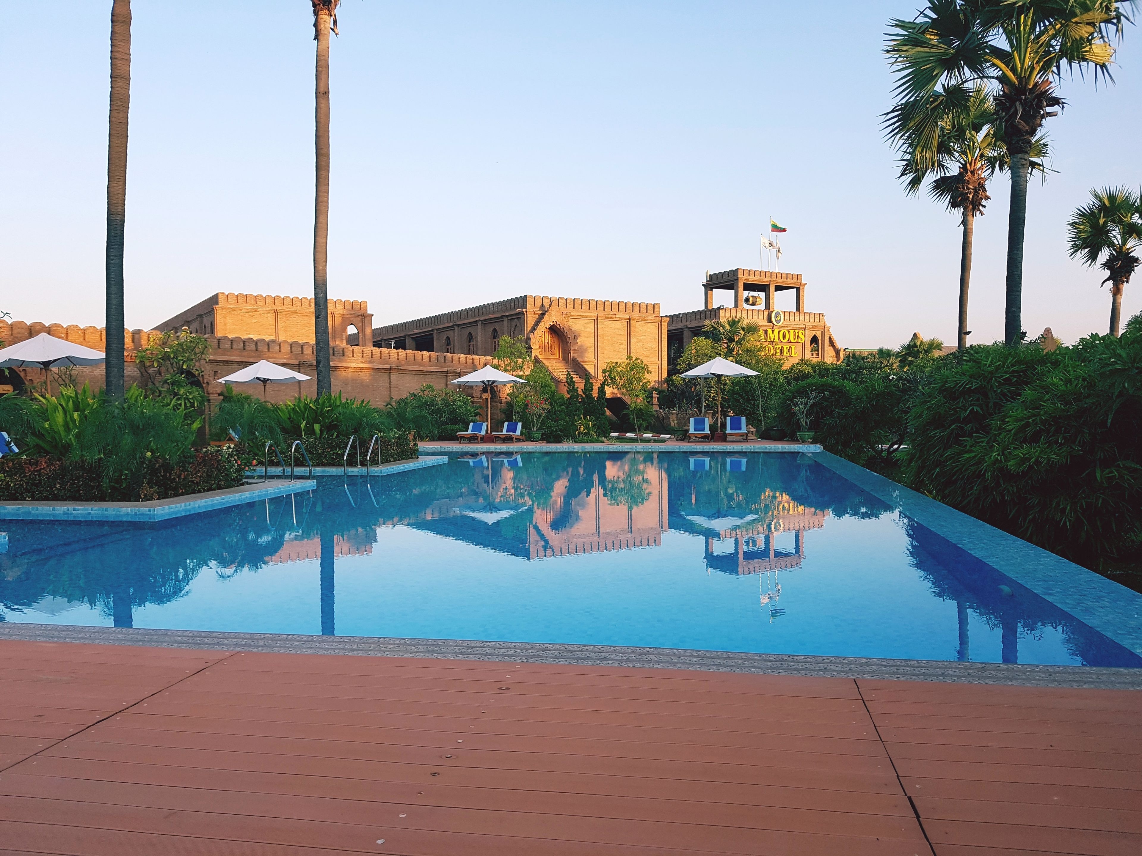 A pool with lounge chairs and umbrellas, surrounded by greenery and a historic building with a flag.