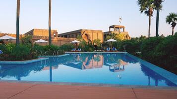 A pool with lounge chairs and umbrellas, surrounded by greenery and a historic building with a flag.