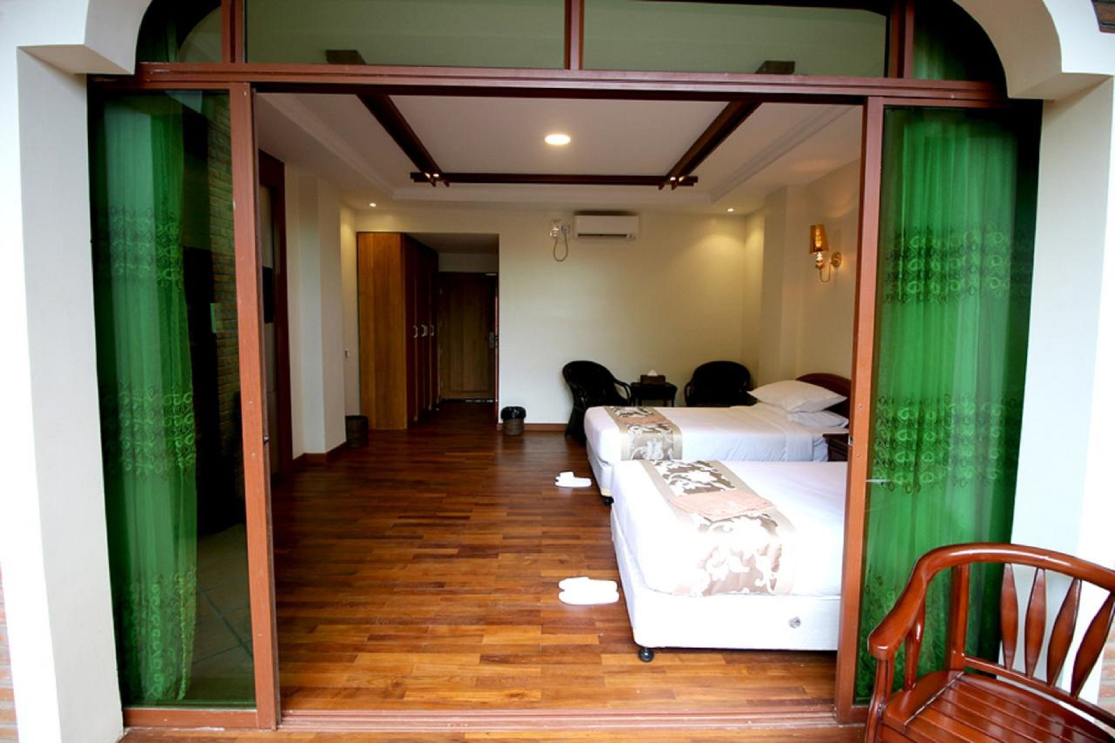 A hotel room with a wooden floor, a bed with a white bedspread and a throw blanket, a wooden chair, and a green curtain.