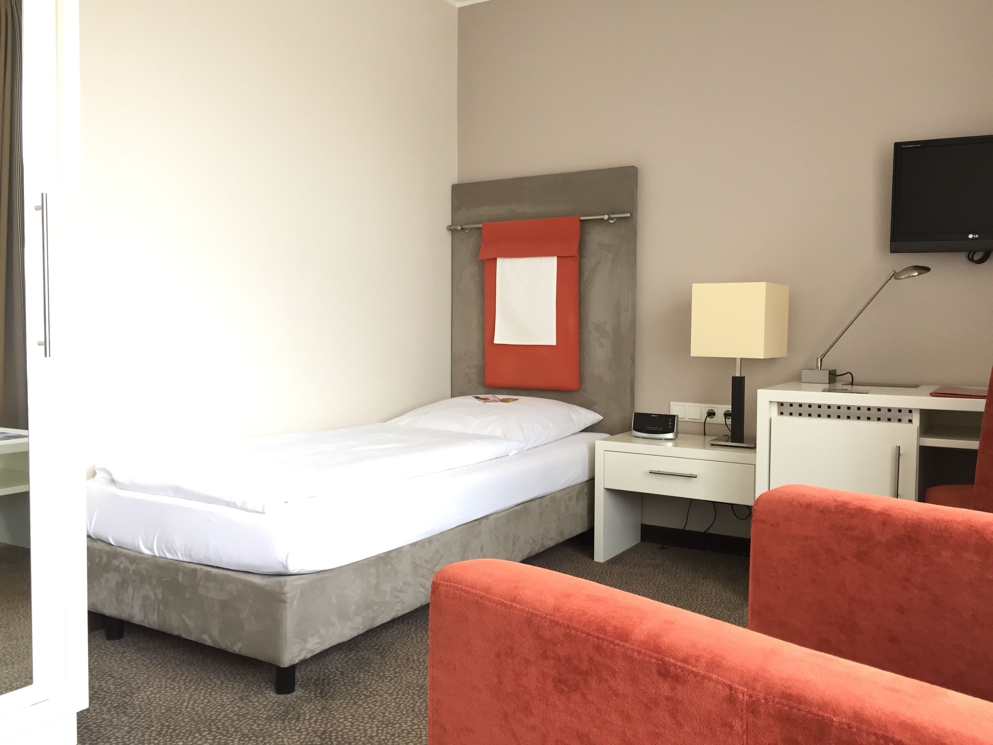 A hotel room with a bed, a desk with a lamp, a television, and a red chair.